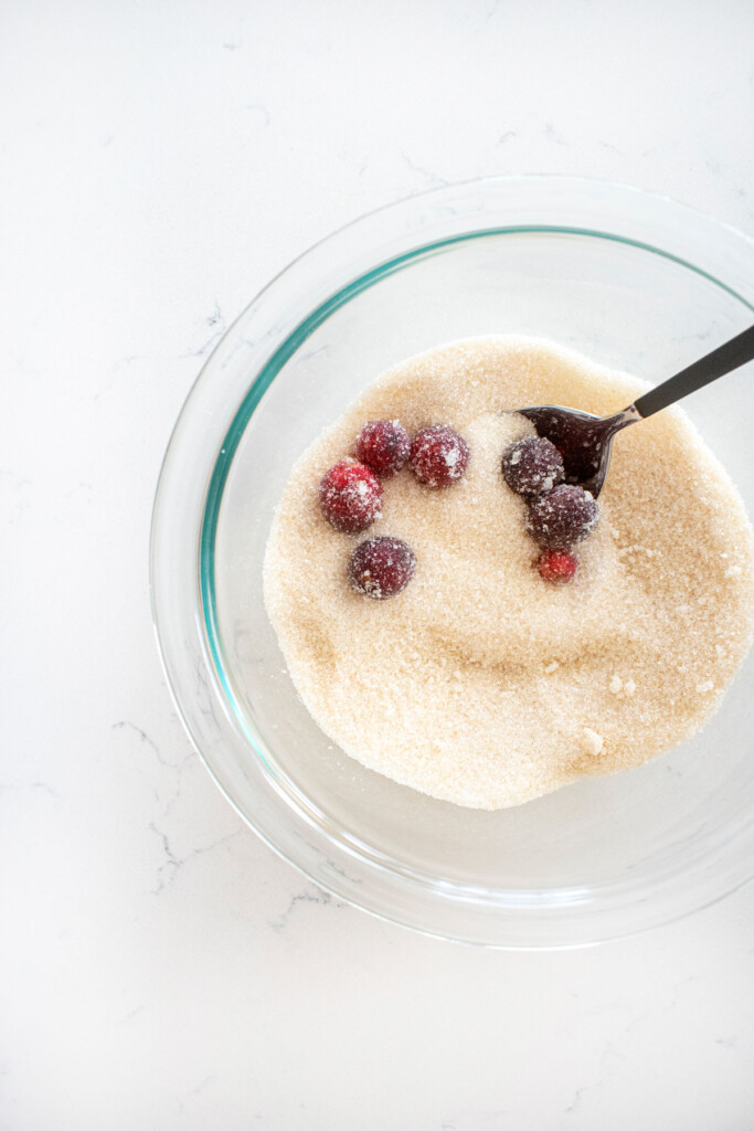 cranberries being coated in a bowl of cane sugar on a white marble counter.