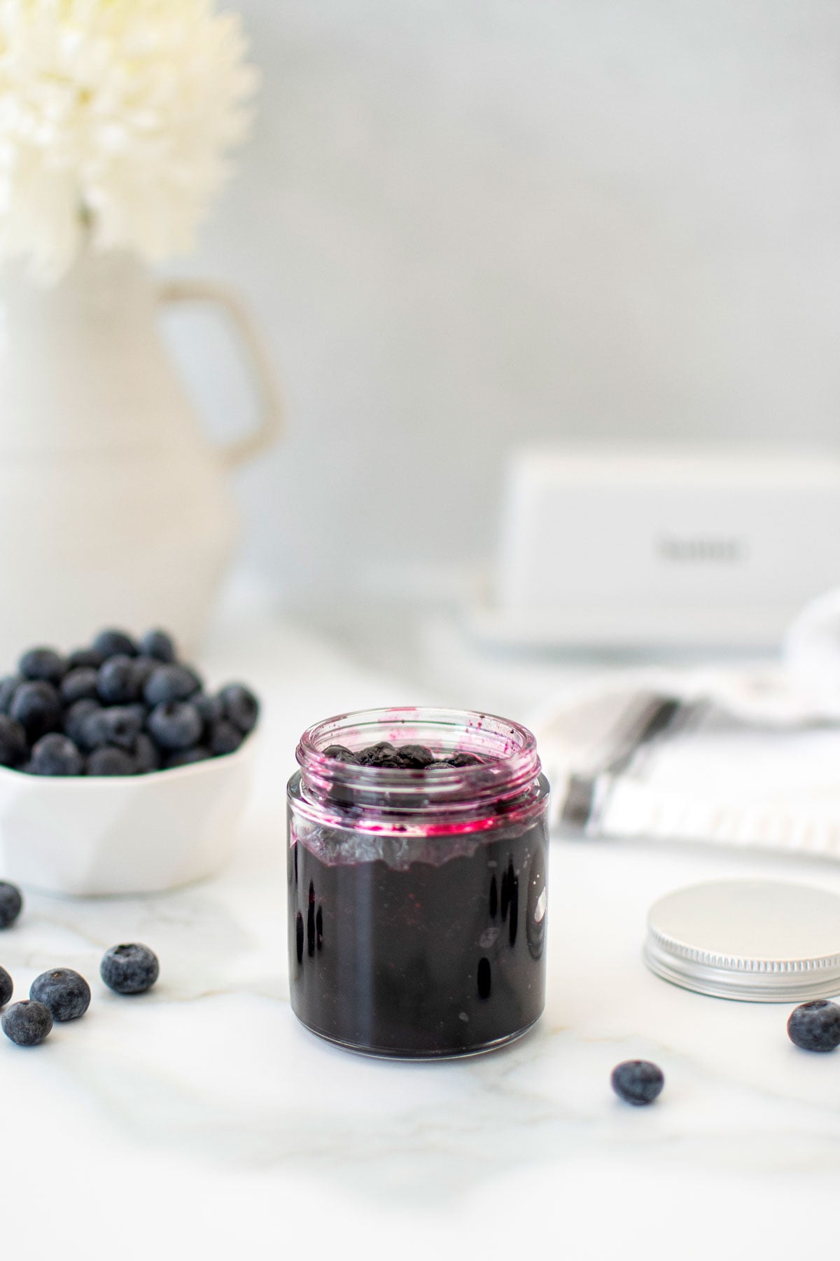 glass jar of homemade blueberry preserves on a white marble counter.