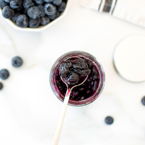 homemade blueberry preserves in a glass jar with a spoon on a white marble counter.