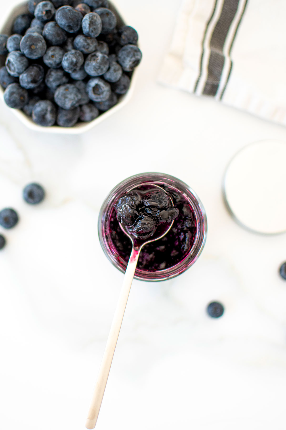 homemade blueberry preserves in a glass jar with a spoon on a white marble counter.