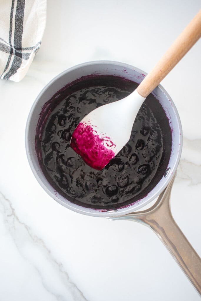 homemade blueberry preserves in a small pot with a spatula on a white marble counter.