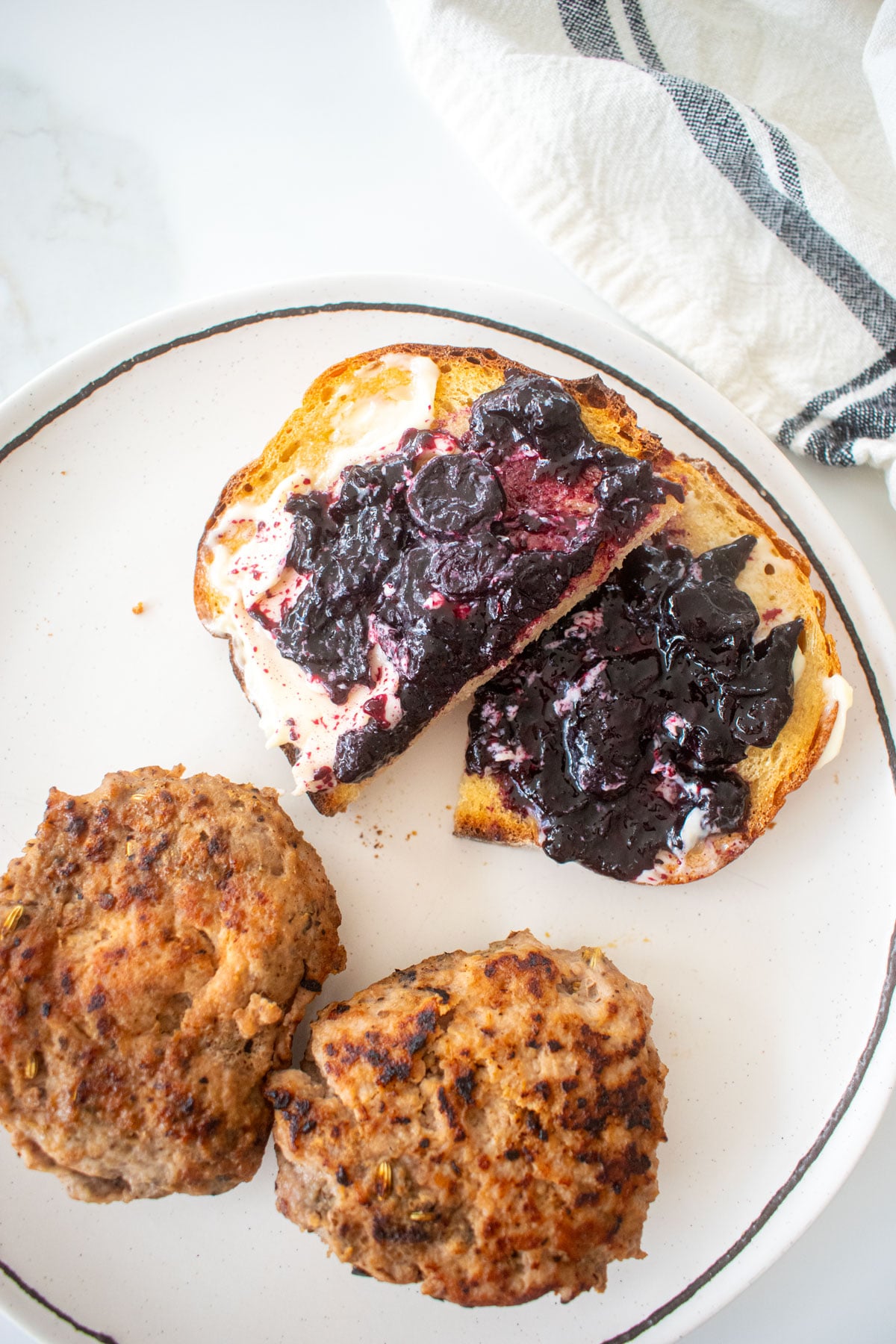 sourdough toast with homemade blueberry preserves and butter on top cut in half on a plate with breakfast sausage on a marble counter.