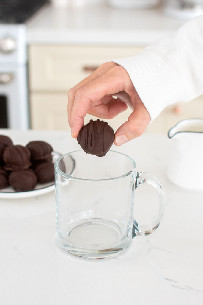woman dropping hot chocolate bomb into a glass mug at kitchen counter.