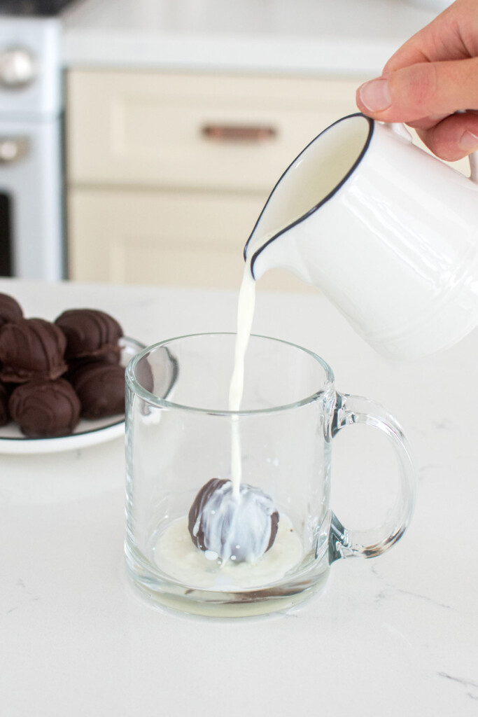 hot chocolate bomb in a glass mug with milk being poured over the top at the kitchen counter.