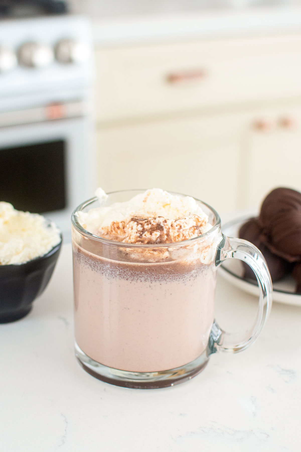 hot cocoa with whipped cream in a glass mug on kitchen counter with hot chocolate bombs next to it.