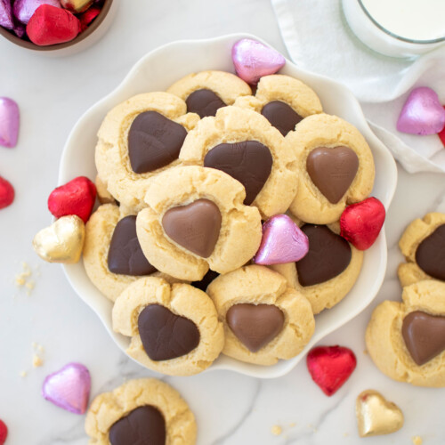 chocolate heart thumbprint cookies on a plate with chocolate wrapped hearts around and a glass of milk on a white marble counter.