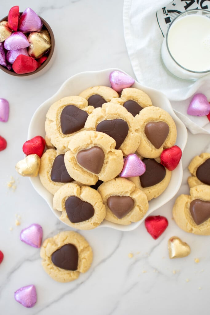 chocolate heart thumbprint cookies on a plate with chocolate wrapped hearts around and a glass of milk on a white marble counter.