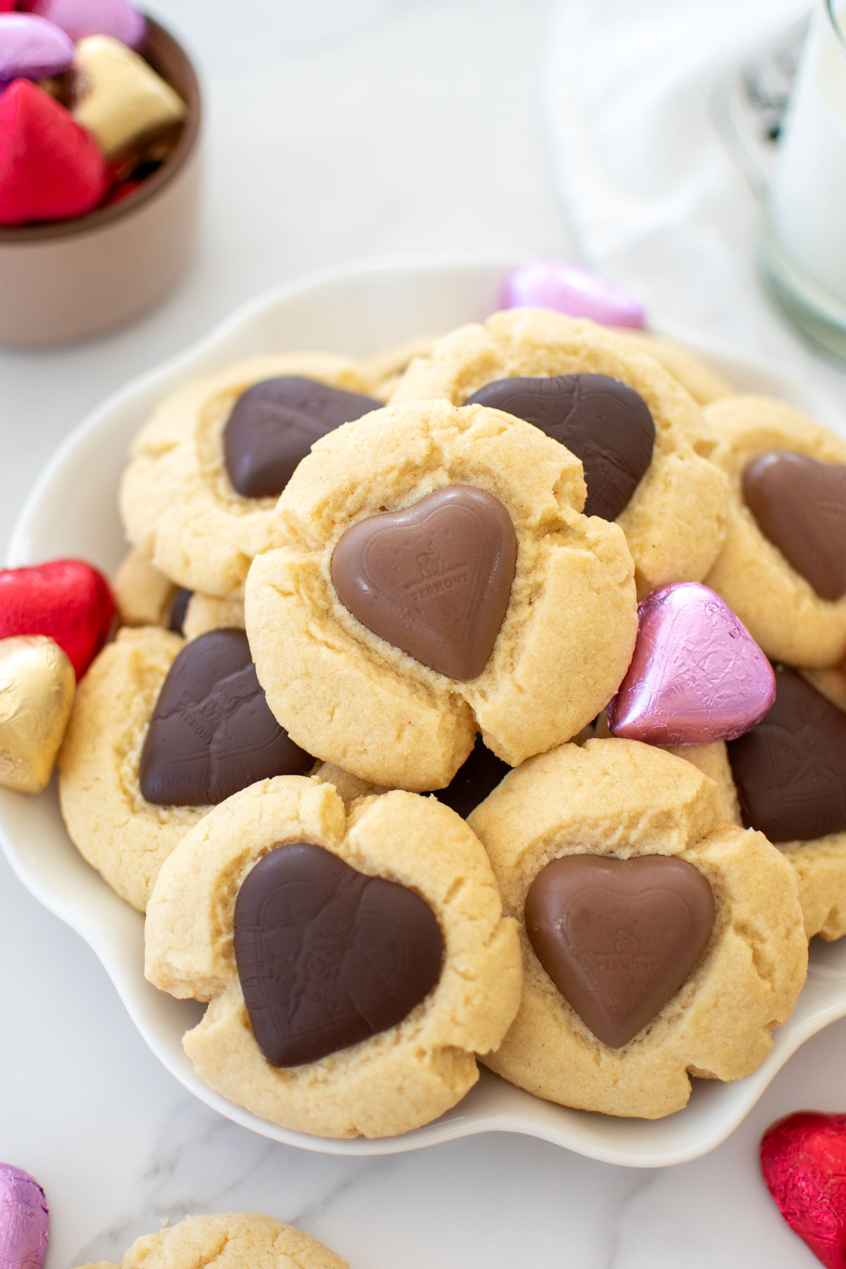 chocolate heart thumbprint cookies on a plate with chocolate wrapped hearts around and a glass of milk on a white marble counter.