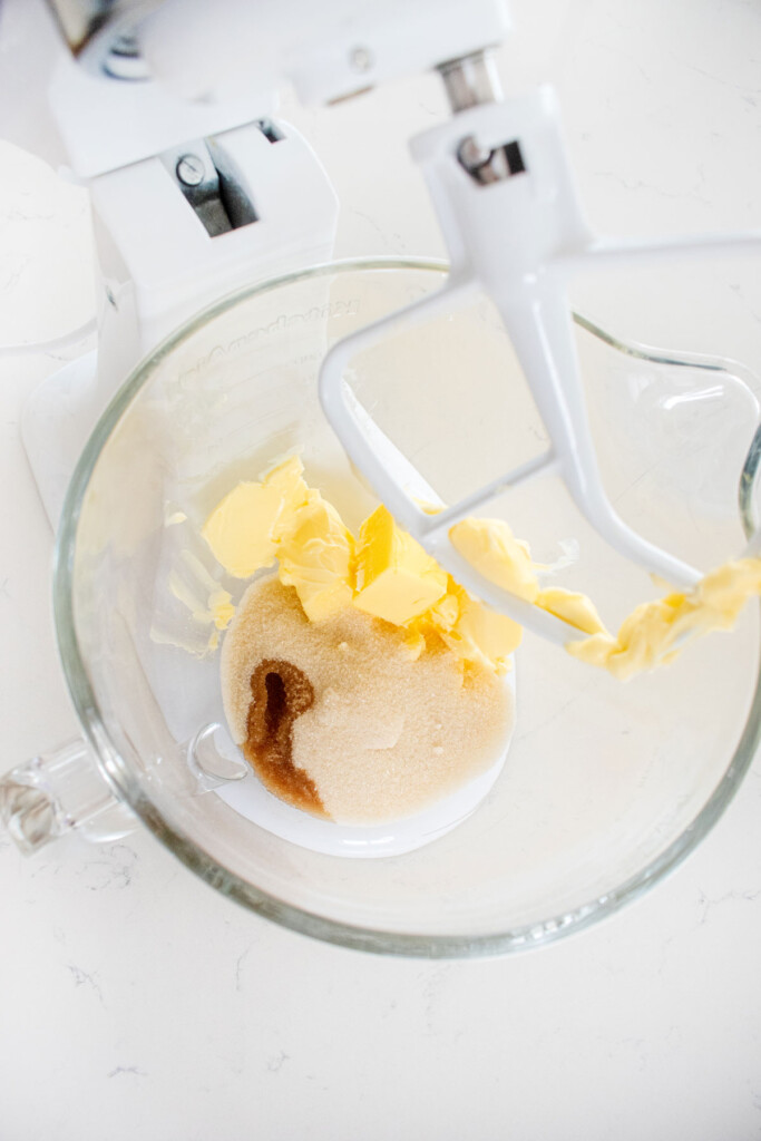 butter, cane sugar and vanilla extract in a glass stand mixing bowl on a white marble counter.