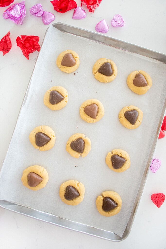 shortbread cookie dough balls pressed with chocolate hearts on a baking sheet on a white marble counter.