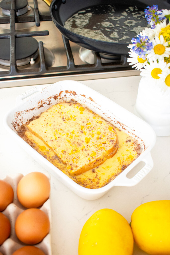 bread soaking in meyer lemon french toast mixture in a white baking dish on the kitchen counter.
