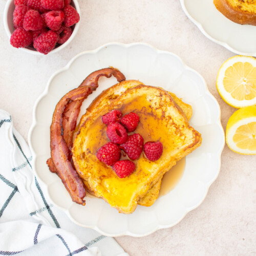 two slices of meyer lemon french toast with raspberries, maple syrup and bacon on a ruffle plate on a breakfast table.
