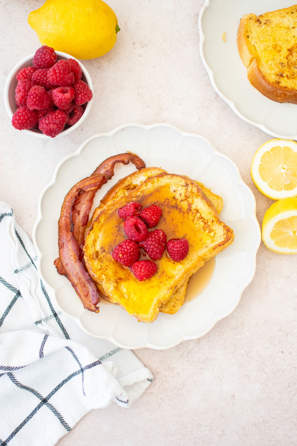 two slices of meyer lemon french toast with raspberries, maple syrup and bacon on a ruffle plate on a breakfast table.