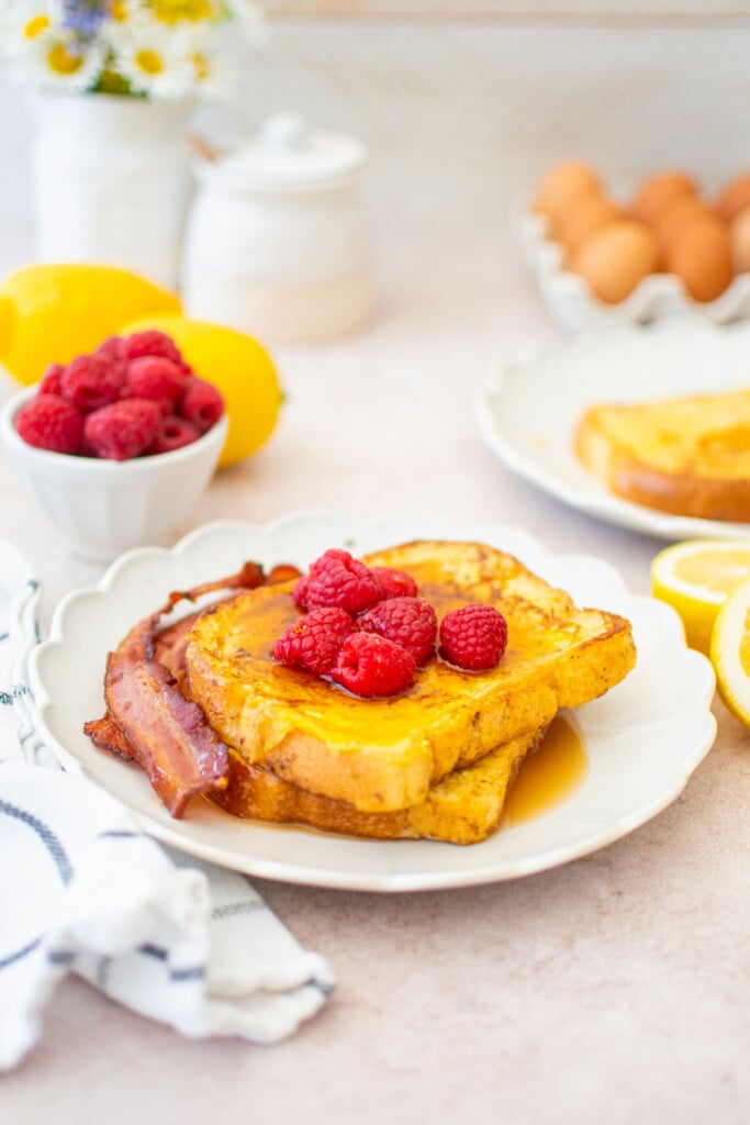 two slices of meyer lemon french toast with raspberries, maple syrup and bacon on a ruffle plate on a breakfast table with fresh flowers.