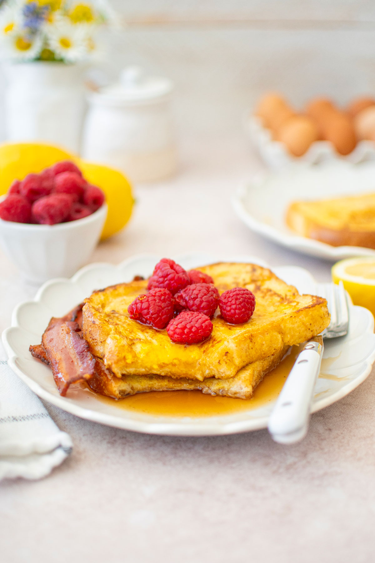 two slices of meyer lemon french toast with raspberries, maple syrup and bacon on a ruffle plate on a breakfast table with fresh flowers.