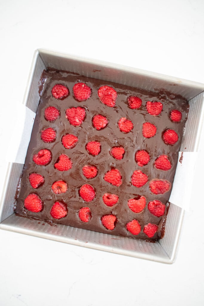 olive oil brownie batter in a baking pan with fresh raspberries on top on a white marble counter.