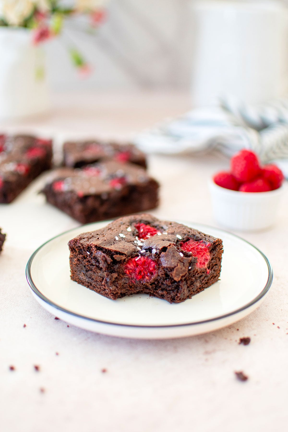 olive oil raspberry brownie on a white plate with fresh flowers and more brownies in the background.