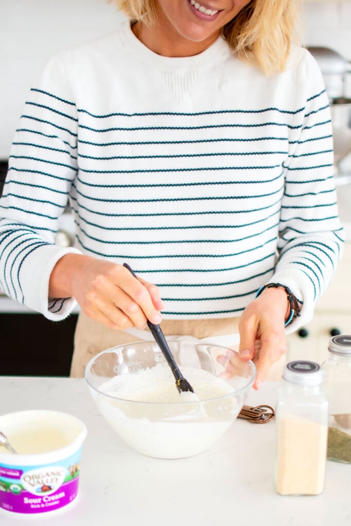 woman wearing an apron mixing up organic dill dip in a glass mixing bowl with a spatula at the kitchen counter.