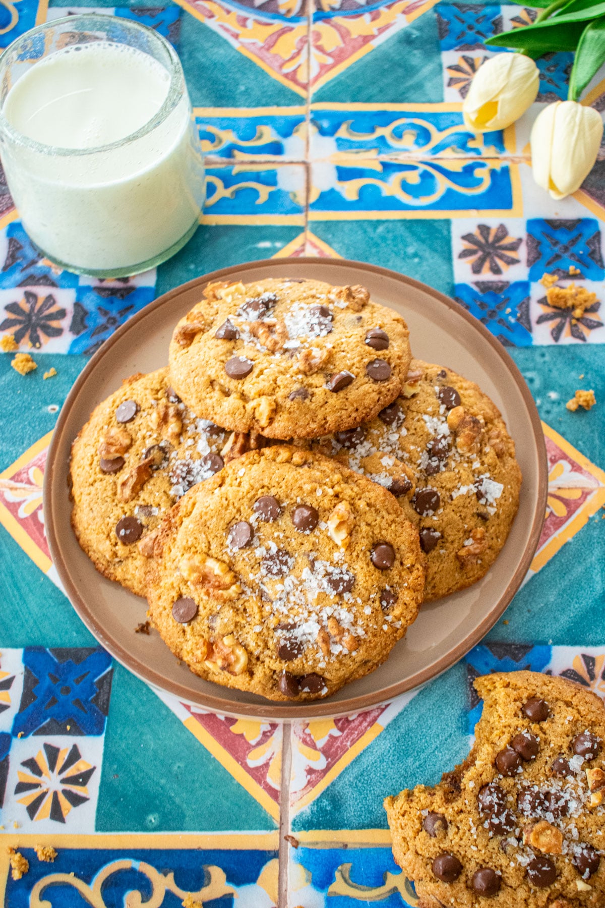 plate of kamut flour chocolate chip cookies on a colorful tile table with a glass of milk and tulips.