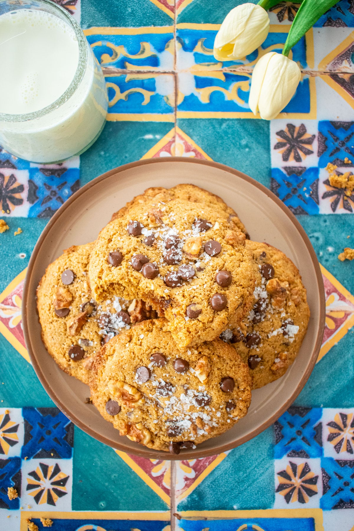 plate of kamut flour chocolate chip cookies on a colorful tile table with a glass of milk and tulips.
