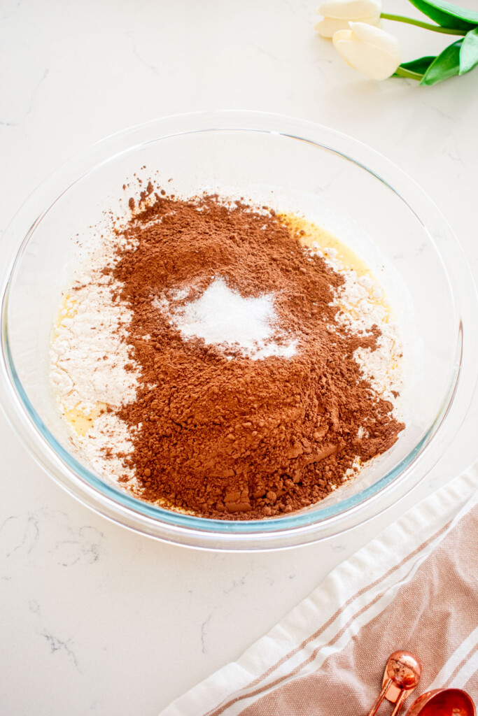 glass mixing bowl with dry ingredients being added to wet ingredients for chocolate cake batter on a white marble counter.