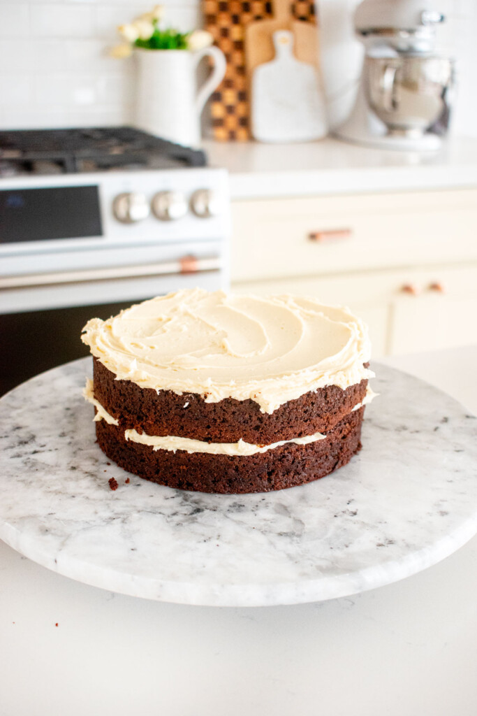 two chocolate cake layers stacked on a white marble cake spinner with vanilla frosting spread on top.