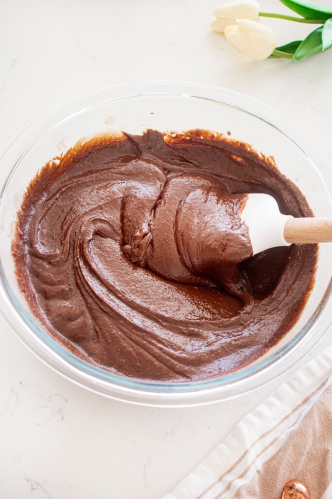 chocolate cake batter in a glass mixing bowl on a white marble counter with a spatula in it.