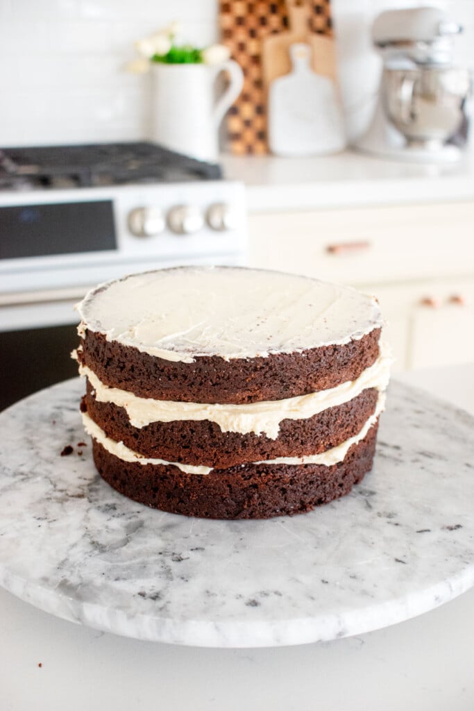 three layer chocolate 6-inch cake being stacked with vanilla frosting on a marble cake spinner on a kitchen counterl