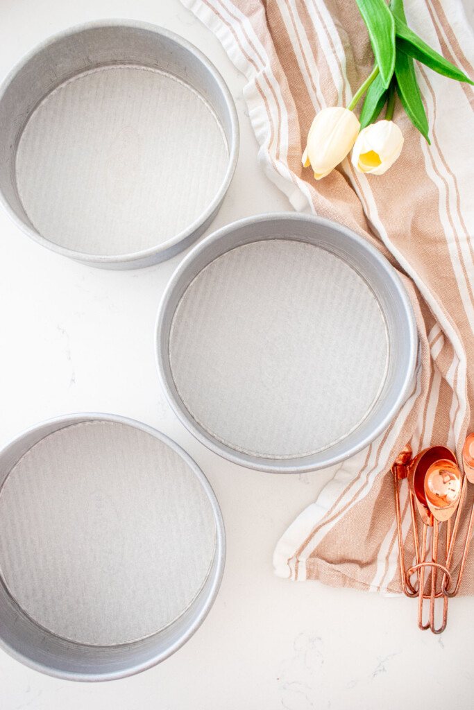 three 6-inch cake pans lined with parchment paper on a white marble counter with tulips and measuring cups.