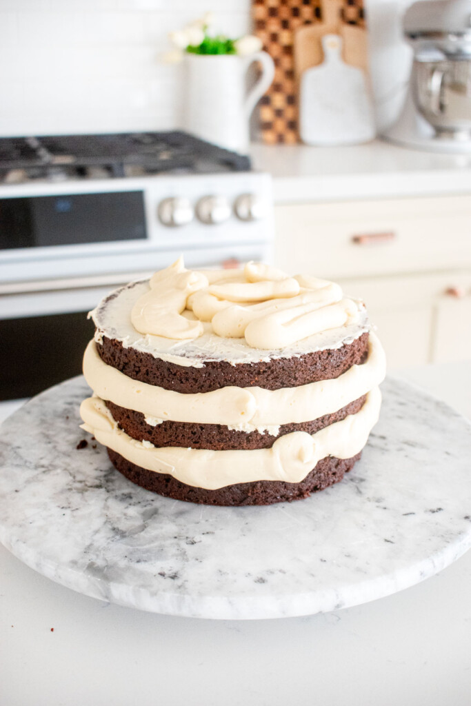 three layer chocolate 6-inch cake being stacked with vanilla frosting on a marble cake spinner on a kitchen counterl