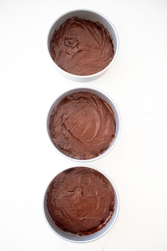 chocolate cake batter in three 6-inch cake pans on a white marble counter.