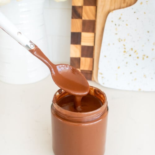 vegan dark chocolate ganache sauce in a glass jar with a spoon spooning some out on a white marble counter.