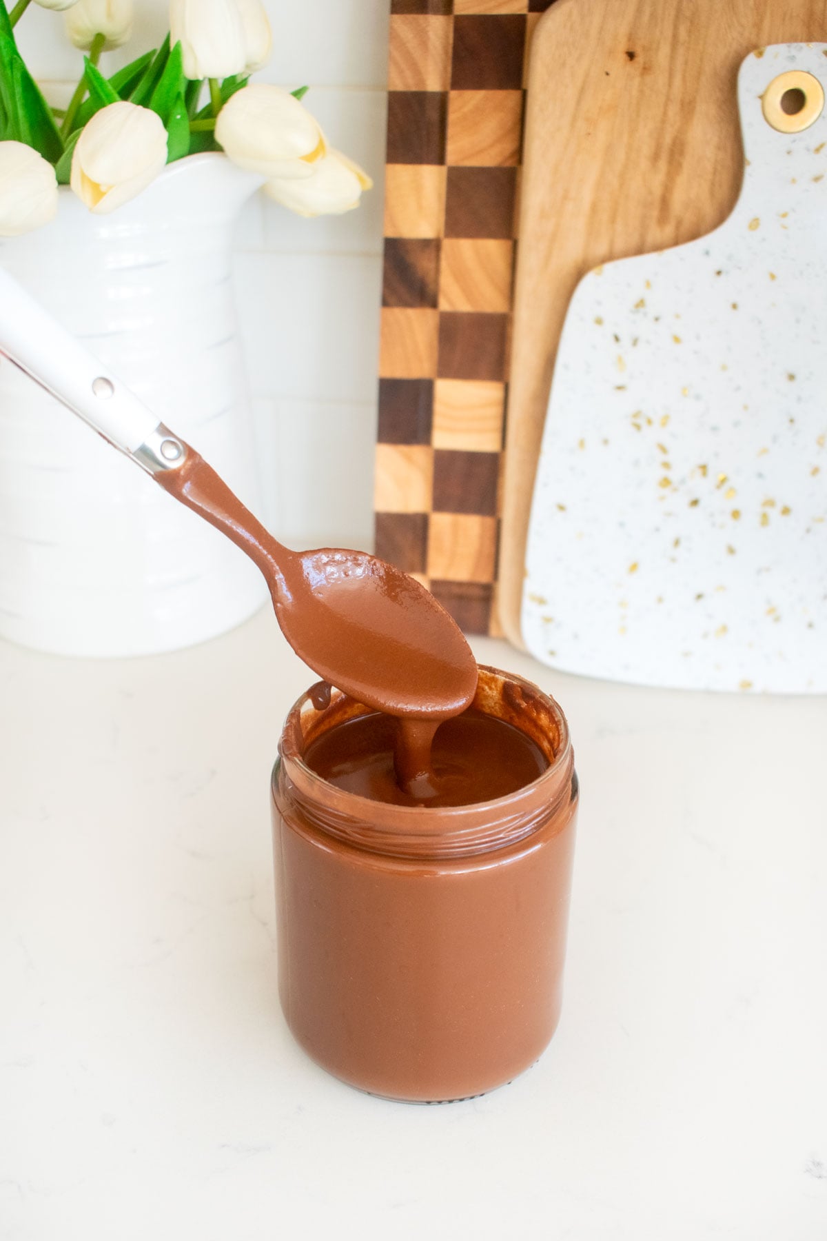vegan dark chocolate ganache sauce in a glass jar with a spoon spooning some out on a white marble counter.