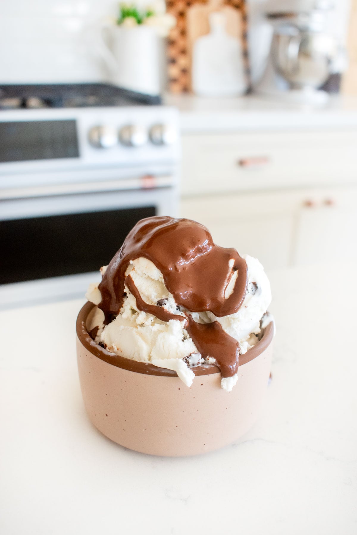 small bowl of chocolate chip ice cream with vegan dark chocolate ganache sauce on a white marble counter.