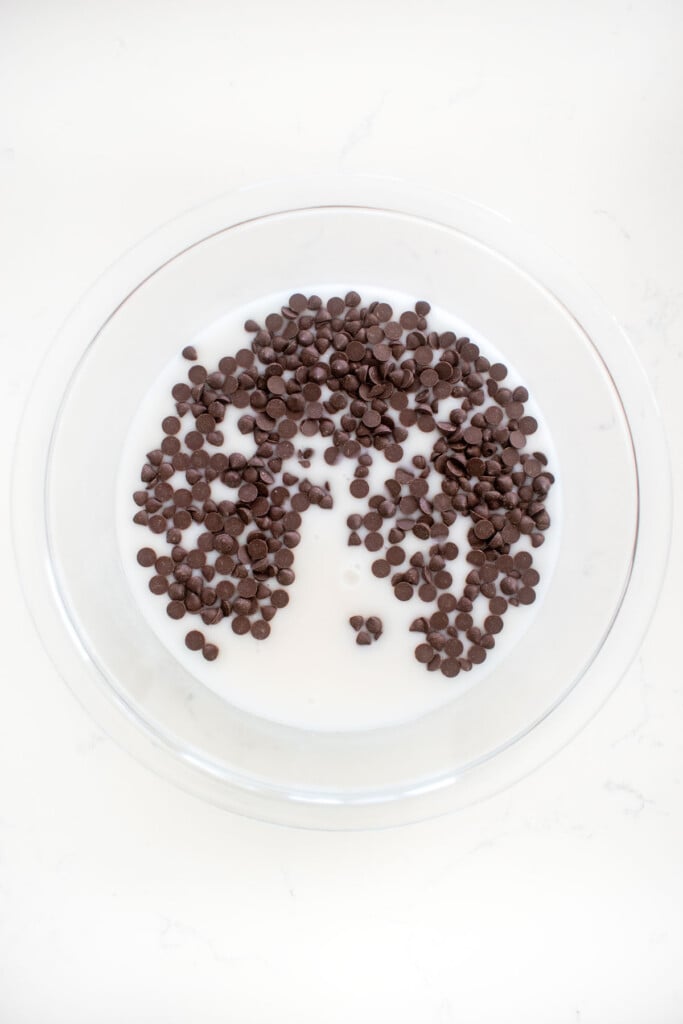 mini dark chocolate chips and coconut milk in a glass mixing bowl on a white marble counter.