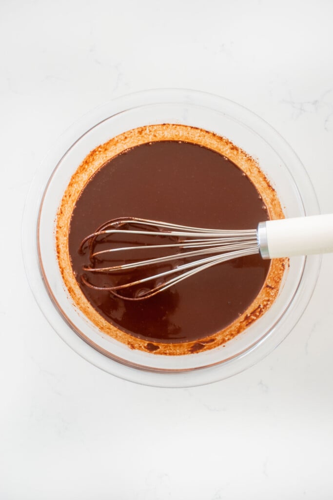 vegan dark chocolate ganache sauce in a glass mixing bowl with a whisk on a white marble counter.