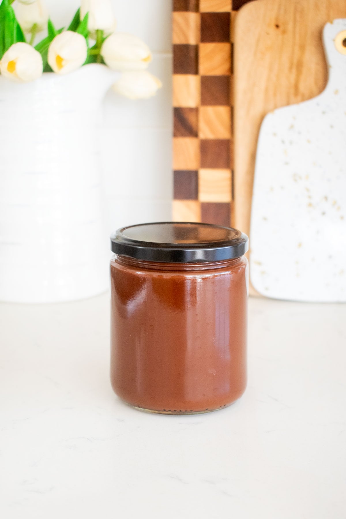 vegan dark chocolate ganache sauce in a glass jar on a white marble counter.