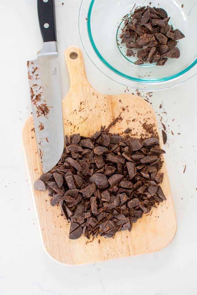 dark chocolate being chopped on a wood cutting board on a white marble counter.
