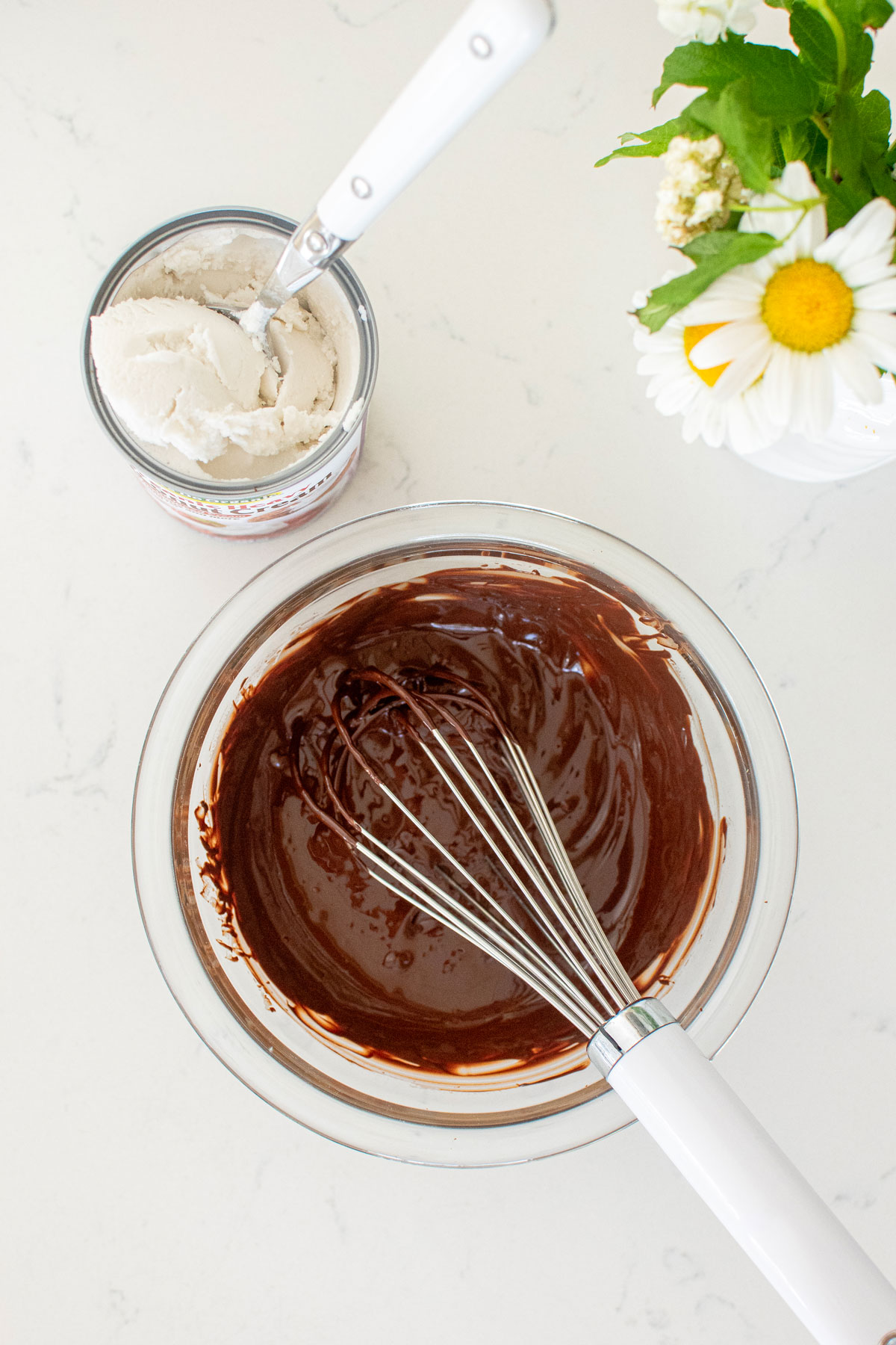 vegan dark chocolate ganache in a glass mixing bowl with a whisk on a white marble counter with coconut cream and flowers on the side.
