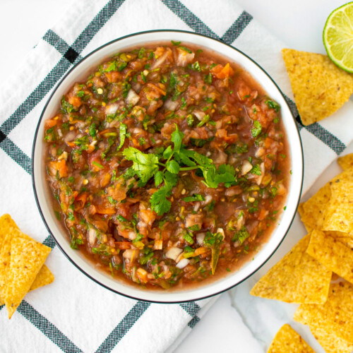 homemade salsa with fresh tomatoes in a bowl with cilantro on top and chips around on a white marble countertop.