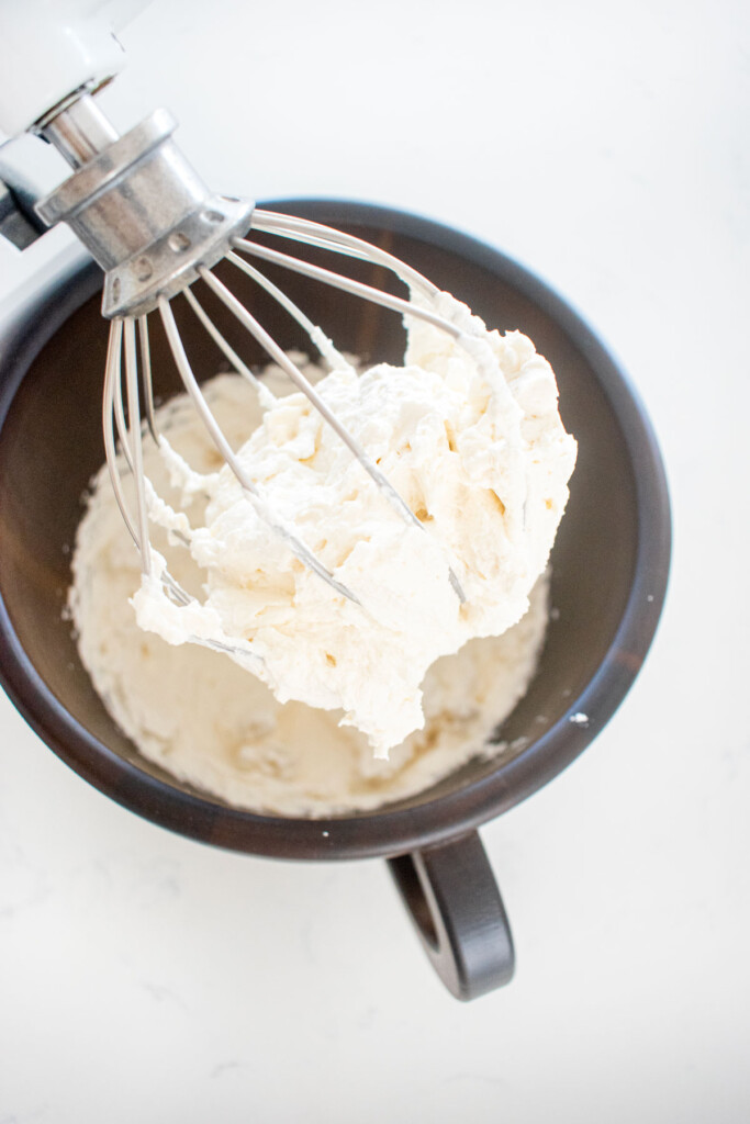 whipped cream frosting on a whisk in a wood mixing bowl on a white marble counter.