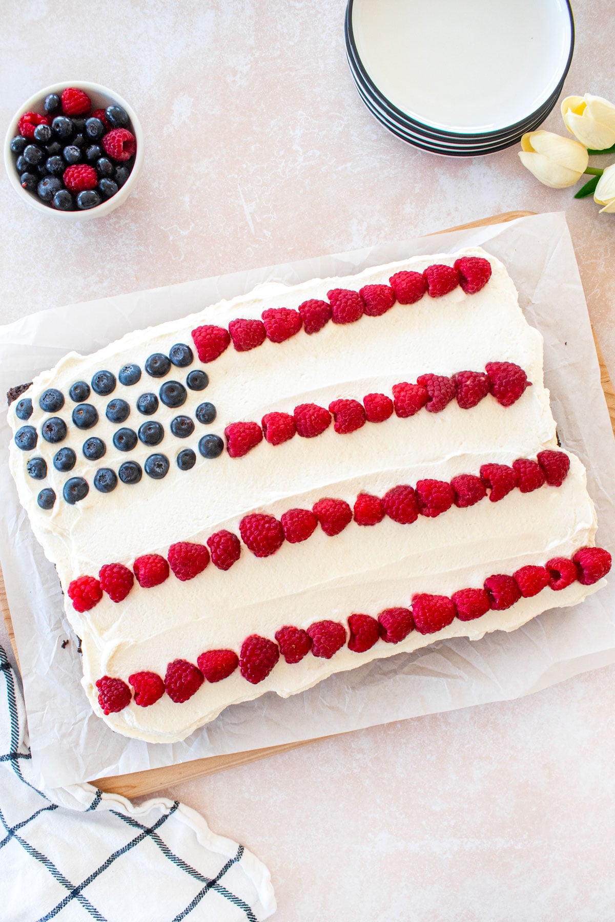 chocolate berry flag cake with whipped cream frosting, raspberries and blueberries in a flag design on a table with berries and dessert plates.