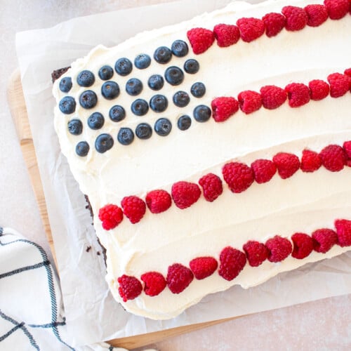chocolate berry flag cake with whipped cream frosting, raspberries and blueberries in a flag design on a table with berries and flowers.