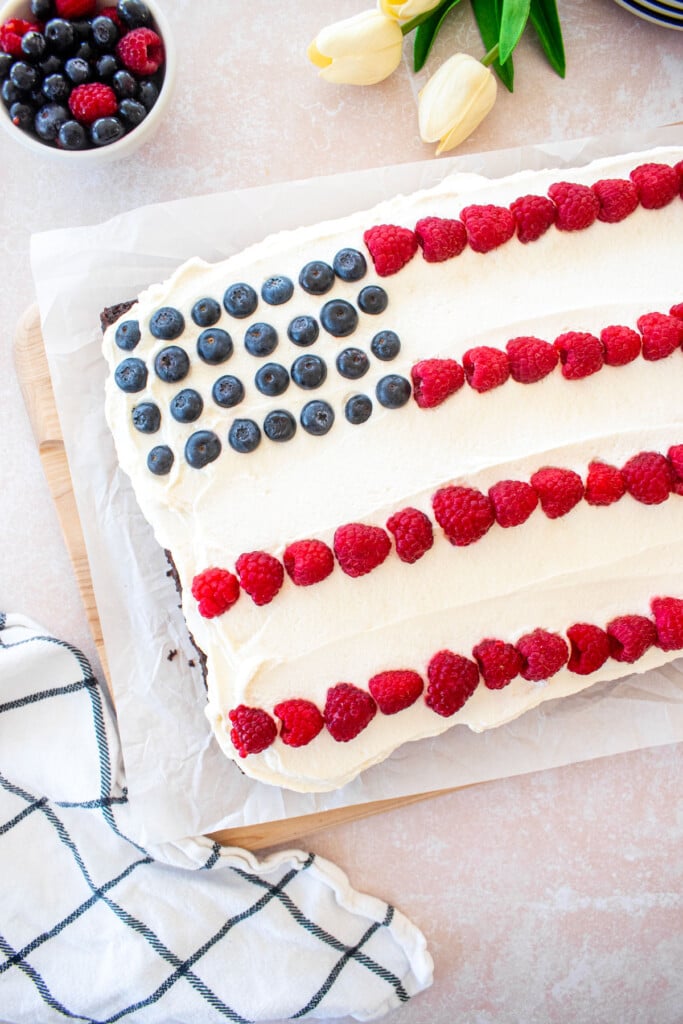 chocolate berry flag cake with whipped cream frosting, raspberries and blueberries in a flag design on a table with berries and flowers.