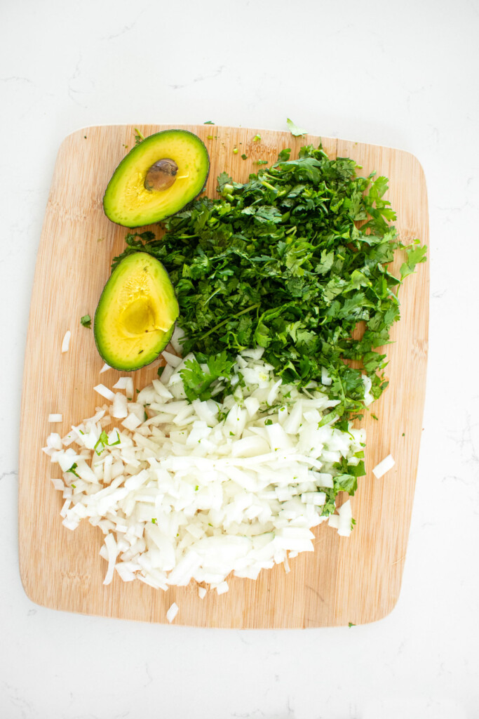 diced onion, chopped cilantro and avocado on a wood cutting board on a marble countertop.