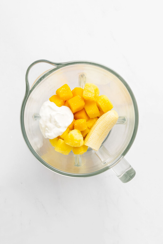 mango, pineapple, banana and greek yogurt in a glass blender on a white marble countertop.