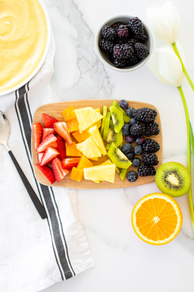 rainbow fruits spread out onto a wood cutting board on a white marble countertop.