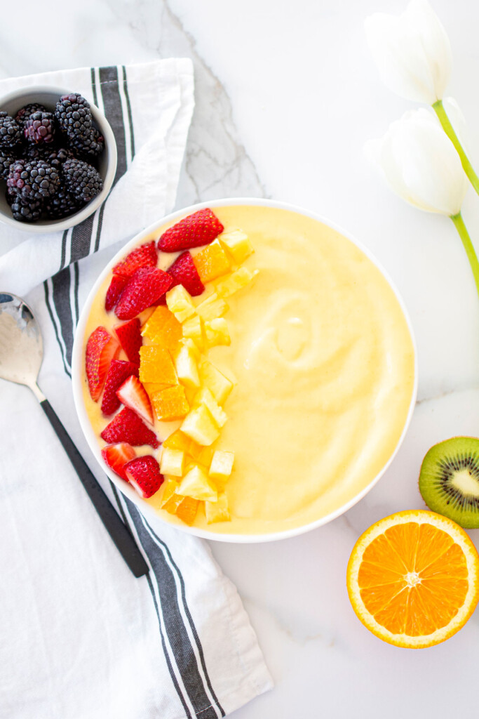 mango smoothie bowl with strawberries, oranges, and pineapple on a white marble countertop.