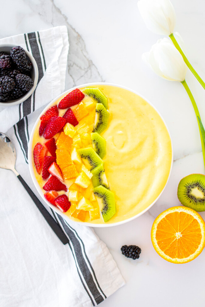 mango smoothie bowl with strawberries, oranges, and pineapple and kiwi on a white marble countertop.