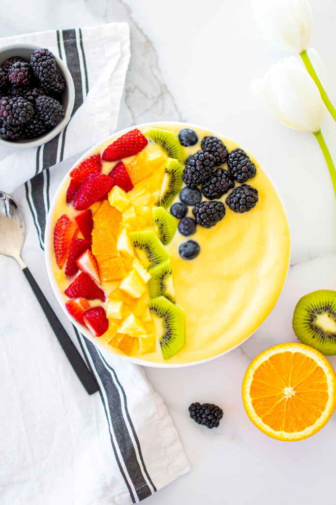rainbow smoothie bowl on a white marble countertop with fresh fruit around.